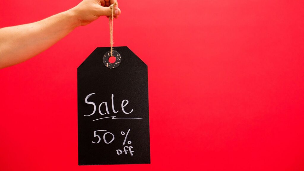 Hand holding a black chalkboard tag reading Sale 50% off against a red background
